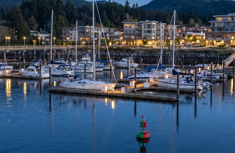 Generated scene: a west-coast harbour at blue hour with a floating marina, a channel buoy and a lit waterfront below forested hills; not a photograph of Nanaimo or the casino.